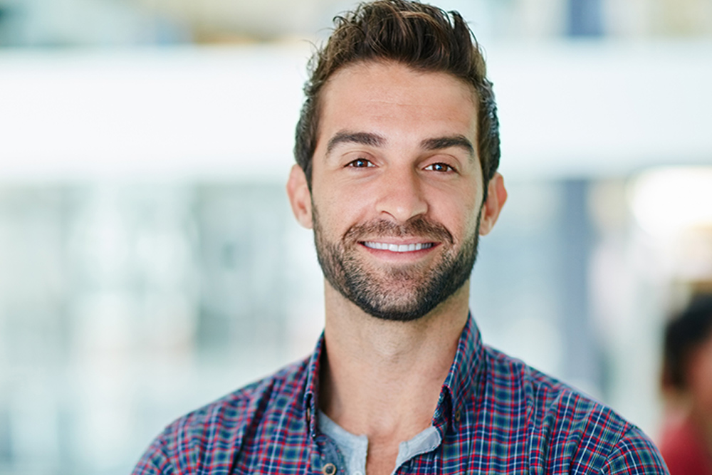 Hombre de aproximadamente 45 años sonriendo. Su cabello castaño es largo y su barba está recortada. Usa una camisa de cuadros