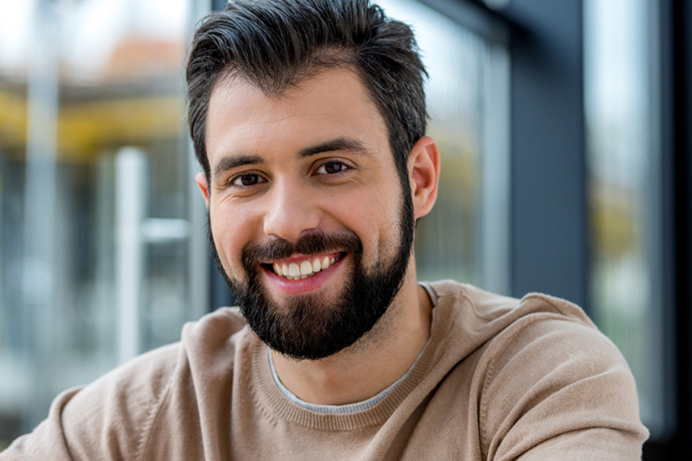 Hombre de aproximadamente 30 años sonriendo. Su cabello es largo y oscuro, al igual que la barba. Está usando un suéter café