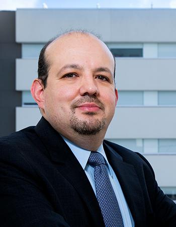 Es la foto de perfil de un cirujano general en Ciudad de México de Hospital Angeles Clínica Londres. El doctor tiene el cabello corto y oscuro, con una frente despejada. Lleva barba y bigote recortados. Está vestido con un traje oscuro, camisa azul clara y una corbata azul con un patrón sutil. El fondo muestra una parte del edificio moderno del hospital, con una fachada de líneas limpias y ventanas amplias.