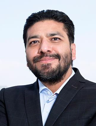 Es la foto de perfil de un ortopedista y traumatólogo en Ciudad de México de Hospital Angeles Clínica Londres. El doctor tiene el cabello oscuro, barba completa y una ligera sonrisa. Está vestido con un traje oscuro y una camisa azul clara. El fondo muestra un cielo despejado, lo que resalta la imagen del doctor.