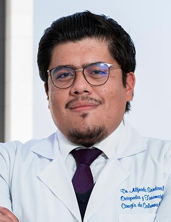 Es la foto de perfil de un ortopedista y traumatólogo en Ciudad de México de Hospital Angeles Clínica Londres. El doctor tiene el cabello oscuro, peinado hacia atrás, y usa gafas. Lleva barba y bigote recortados. Está sonriendo ligeramente y viste una bata médica blanca con su nombre y la especialidad 'Ortopedia y Traumatología, Cirugía de Columna' bordados en el pecho. Lleva una corbata color púrpura oscuro. El fondo muestra una parte del edificio moderno del hospital.