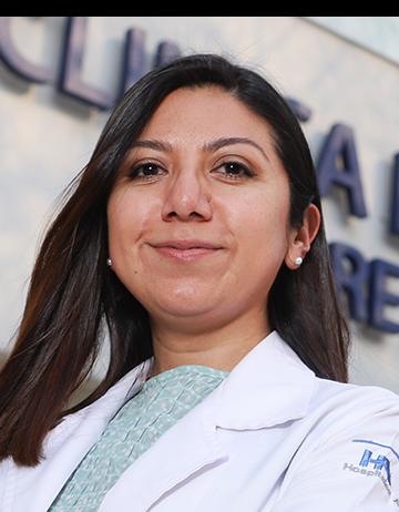 Es la foto de perfil de una internista en Ciudad de México de Hospital Angeles Clínica Londres. La doctora tiene el cabello oscuro y lacio, con una expresión tranquila. Viste una bata médica blanca sobre una blusa de color verde claro. En la manga de la bata se distingue el logotipo del Hospital Ángeles. El fondo muestra un edificio del hospital con un letrero en letras grandes.