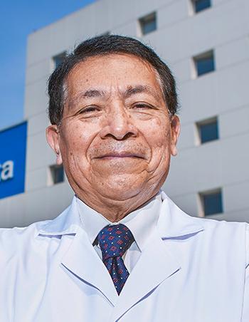 Es la foto de perfil de un ortopedista y traumatólogo en Ciudad de México de Hospital Angeles Clínica Londres. El doctor tiene el cabello canoso y corto, y lleva una expresión amigable con una leve sonrisa. Viste una bata médica blanca y una corbata azul con un patrón discreto. El fondo muestra un edificio del hospital con un cielo despejado y ventanas en la fachada.