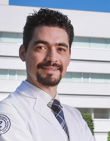 Es la foto de perfil de un cirujano plástico en Ciudad de México de Hospital Angeles. El doctor viste una bata blanca con una camisa blanca y una corbata de color beige y azul marino. Tiene barba de candado, cejas pobladas y cabello rizado.