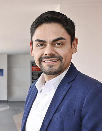 Es la foto de perfil de un oncólogo médico en León de Hospital Angeles León. Utiliza un saco azul marino, camisa blanca y está en el pasillo del hospital.
