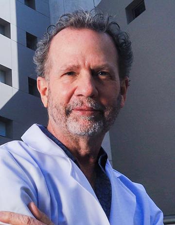 Es la foto de perfil de un ortopedista y traumatólogo en Huixquilucan de Hospital Angeles Lomas. El doctor viste una bata blanca con una playera oscura de fondo, tiene cabello canoso y rizado y barba canosa.