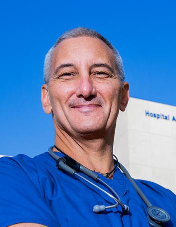Es la foto de perfil de un cirujano general en Huixquilucan de Hospital Angeles Lomas. Usa un uniforme azul, tiene un estetoscopio sobre los hombros, tiene el cabello canoso y está sonriendo.