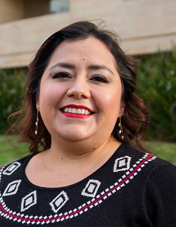 Es la foto de perfil de una oncóloga médica especialista en medicina interna en Ciudad de México de Hospital Angeles. La doctora viste una blusa negra con rombos pequeños en el cuello de color blanco. Tiene aretes largos y está sonriendo. 