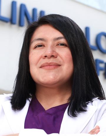 Es la foto de perfil de una gastroenteróloga y endoscopista en Ciudad de México de Hospital Angeles Clínica Londres. La doctora tiene el cabello oscuro, lacio y de longitud media. Viste una bata médica blanca sobre un uniforme de color púrpura y tiene una expresión confiada. El fondo muestra un edificio del hospital con un letrero en letras grandes. 