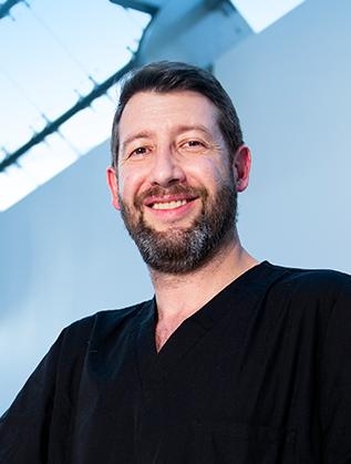Es la foto de perfil de una nutrióloga en Huixquilucan de Hospital Angeles Lomas. Usa un uniforme negro en cuell V, tiene baraba, está sonriendo y es de tez blanca.