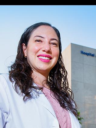 Es la foto de perfil de una ginecóloga y obstetra en Huixquilucan de Hospital Angeles Lomas. Usa una bata blanca, tiene un suéter rosa, cabello rizado y está sonriendo.