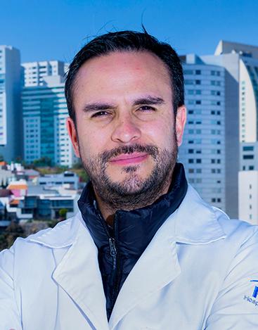 Es la foto de perfil de un neurólogo pediátrico en Huixquilucan de Hospital Angeles Lomas. Usa una bata blanca con un suéter de cuello alto azul marino, es de tez blanca y cabello castaño oscuro. Sus cejas son pobladas y está sonriendo.