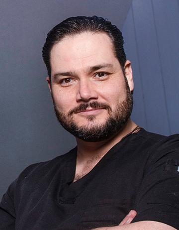 Es la foto de perfil de un otorrinolaringólogo en Huixquilucan de Hospital Angeles Lomas. El doctor tiene barba y bigote oscuro. Tiene un uniforme médico de color negro. 