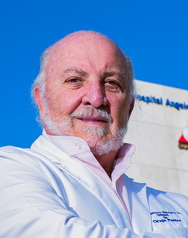 Es la foto de perfil de un cirujano plástico en Huixquilucan de Hospital Angeles Lomas. Usa una bata blanca con una camisa rosa, tiene los brazos cruzados y está sonriendo.