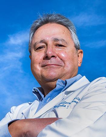 Es la foto de perfil de un ortopedista y traumatólogo en Huixquilucan de Hospital Angeles Lomas. Usa bata blanca con camisa azul, tiene los brazos cruzados y su cabello es canoso.