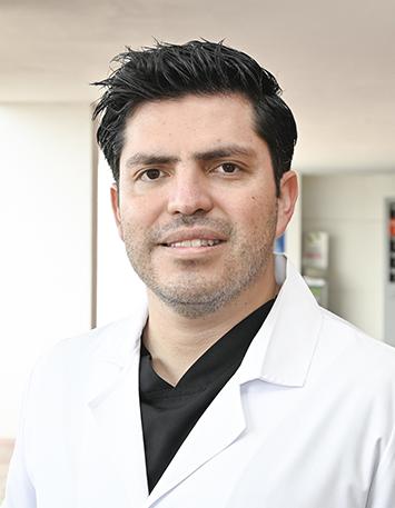 Es la foto de perfil de un cirujano maxilofacial en León de Hospital Angeles León. Utiliza una bata blanca con una playera negra debajo, está sonriendo.