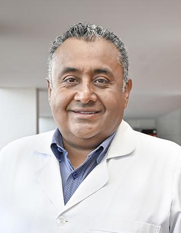 Es la foto de perfil de un pediatra en León de Hospital Angeles León. Utiliza una bata blanca con una camisa azul. El doctor es canoso y está sonriendo. 