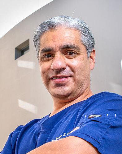 Es la foto de perfil de un cirujano oncológico en Huixquilucan de Hospital Angeles Lomas. El doctor usa un uniforme azul con cuello redondo, tiene cabello canoso y los brazos cruzados.