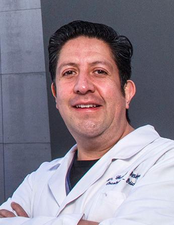 Es la foto de perfil de un oncólogo médico pediátrico en Huixquilucan de Hospital Angeles Lomas. Usa bata blanca, playera negra debajo y está sonriendo.