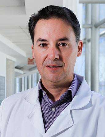 Es la foto de perfil de un cirujano de tórax pediátrico en Huixquilucan de Hospital Angeles Lomas. Tiene una camisa morada, usa bata y de fondo está un pasillo del hospital. 