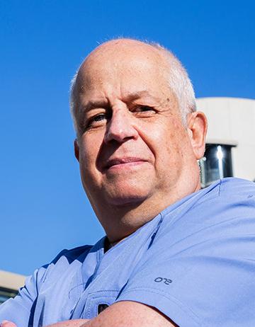 Es la foto de perfil de un médico general en Huixquilucan de Hospital Angeles Lomas. Usa un uniforme azul cielo, tiene cabello canoso y los brazos cruzados.