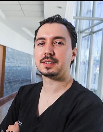 Es la foto de perfil de un cirujano general en Huixquilucan de Hospital Angeles Lomas. está usando un uniforme negro con cuello V. Tiene los brazos cruzados y cabello oscuro.