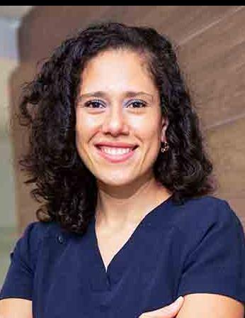 Es la foto de perfil de una gastroenteróloga en Ciudad de México de Hospital Angeles Santa Mónica. Usa un uniforme médico azul. Tiene el cabello chino y de color café oscuro. Está sonriendo y tiene los brazos cruzados.