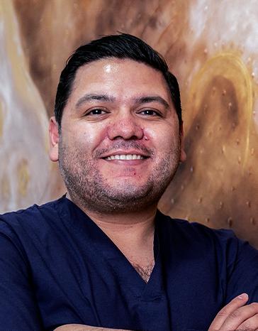 Es la foto de perfil de un urólogo en León de Hospital Angeles León. Utiliza ununiforme en cuello V de color azul marino. Tiene los brazos cruzados y está sonriendo.