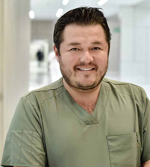 Es la foto de perfil de un oftalmólogo en Querétaro de Hospital Angeles Centro Sur. Viste un uniforme verde olivo, está sonriendo y de fondo se ve el pasillo del hospital.