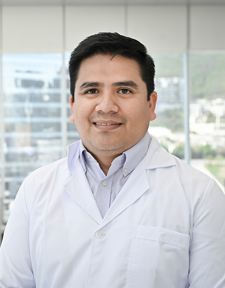 Es la foto de perfil de un cirujano oncólogo en San Pedro Garza García de Hospital Angeles Valle Oriente. Usa una bata médica blanca y una camisa color azul claro con cuadros. Tiene el cabello negro y está sonriendo ligeramente.