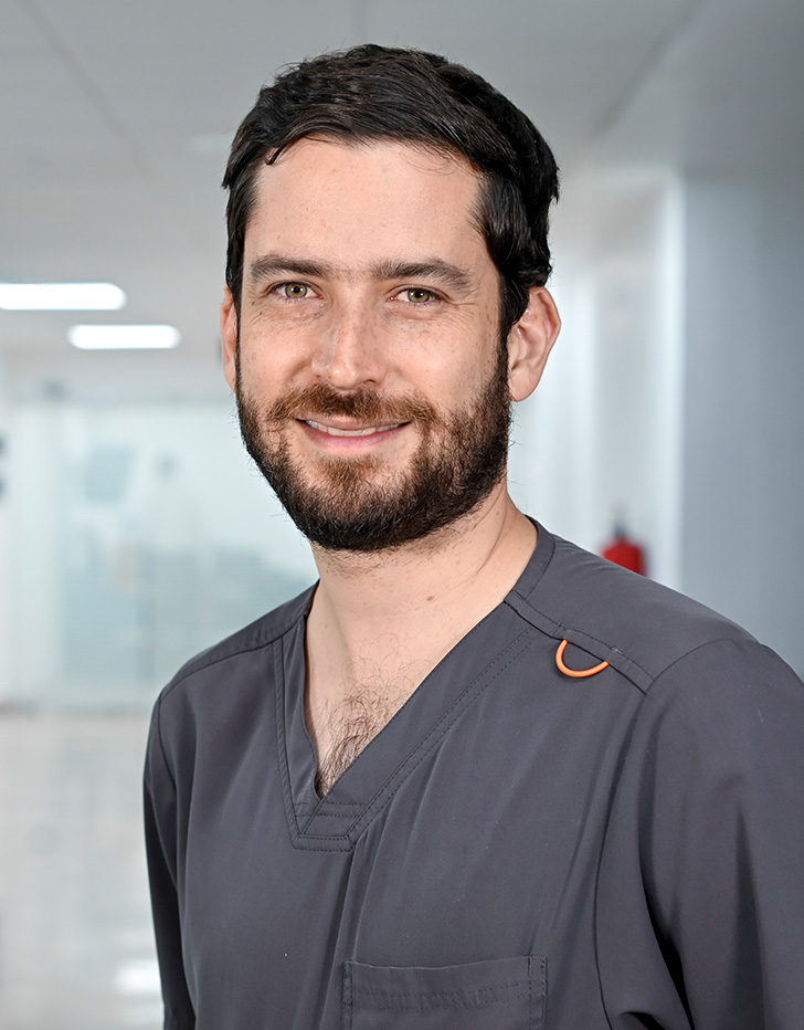 Es la foto de perfil de un angiólogo y cirujano vascular en Guadalajara de Hospital Angeles Del Carmen. Usa un uniforme médico color gris. Tiene el cabello y barba color café oscuro. Tiene los brazos cruzados por detrás.