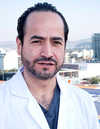 Es la foto de perfil de un urólogo en San Luis Potosí de Hospital Angeles San Luis Potosí. Usa una bata blanca con un uniforme médico azul. Tiene barba y cabello negro.