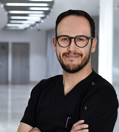 Es la foto de perfil de un cirujano pediátrico en Huixquilucan. Tiene el cabello negro. Usa lentes y un uniforme médico negro. Tiene los brazos cruzados y está sonriendo.