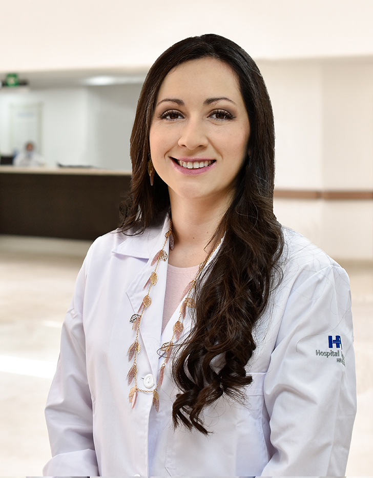 Es la foto de perfil de una cirujana de trasplantes y nefróloga pediátrica en Querétaro de Hospital Angeles Queretaro. Usa una bata color blanco con una blusa color rosa claro. Tiene puesto un collar largo y doorado que tiene representaciones de una hoja. Tiene el cabello largo y ondulado.