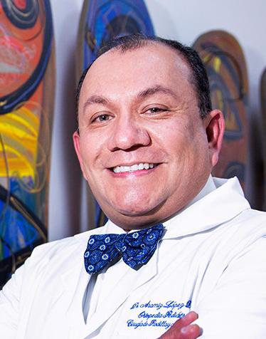 Es la foto de perfil de un ortopedista y traumatólogo pediátrico en Ciudad de México de Hospital Angeles Lindavista. Usa una bata y camisa blanca con un moño azul con estampado.