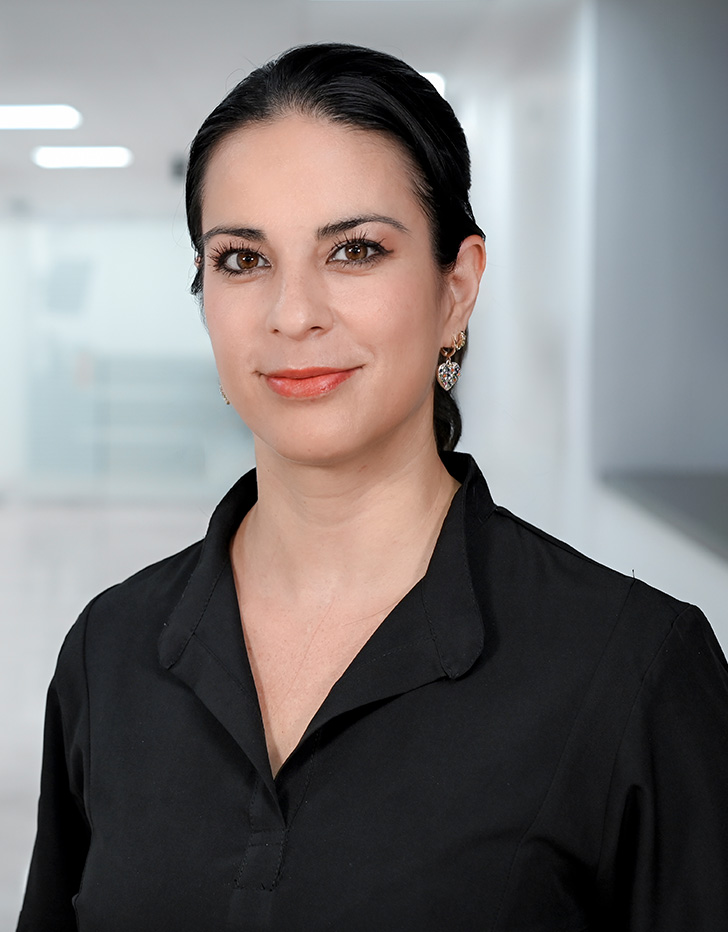 Es la foto de perfil de una otorrinolaringóloga en Guadalajara de Hospital Angeles. Usa una blusa color negra con unos aretes grandes de forma de corazón dorados con otros colores. Tiene el cabello negro y recogido.