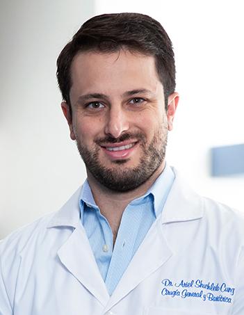 Es la foto de perfil de un cirujano general en Huixquilucan. Tiene el cabello y barba café. Usa una bata blanca con una camisa azul.