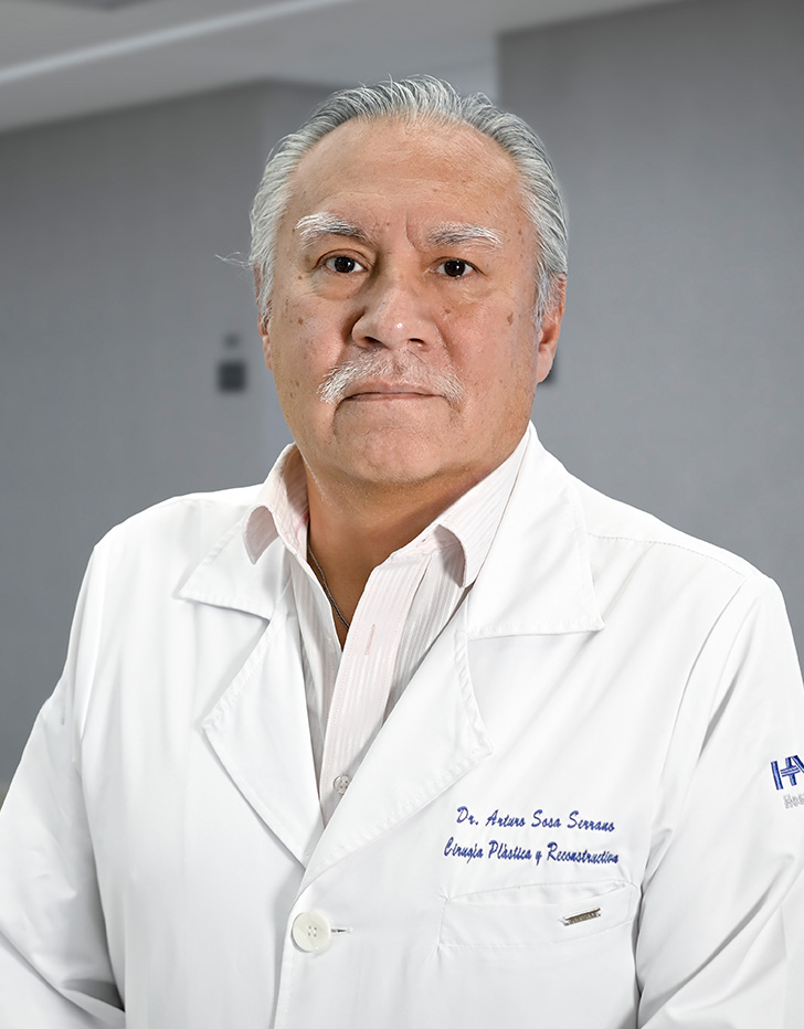 Es la foto de perfil de un cirujano plástico en Ciudad de México de Hospital Angeles Lindavista. Tiene el cabello negro con gris. Usa bata blanca y una camisa de cuadros blanco y verde.