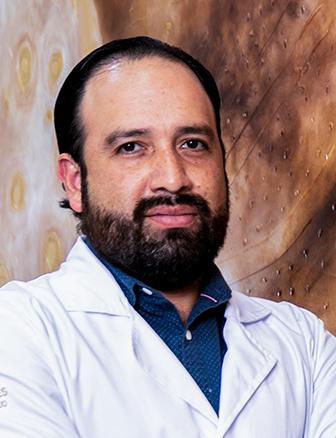 Es la foto de perfil de un cirujano oncológico en Querétaro de Hospital Angeles Centro Sur. Usa una bata blanca, una camisa azul marino con lunares y tiene barba y bigote. Nuestro especialista brinda sus servicios en el Hospital Centro Sur y en León.