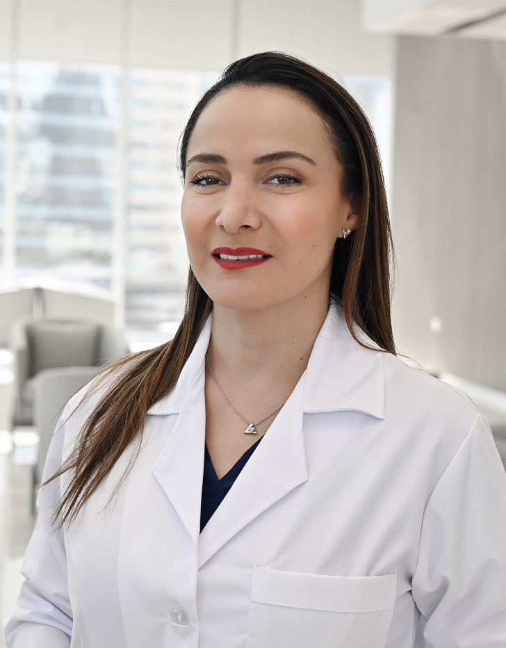 Es la foto de perfil de una bióloga de la reproducción y ginecóloga y obstetra en San Pedro Garza García de Hospital Angeles. Usa una bata blanca con un uniforme médico color azul oscuro. Tiene pusto joyería color plata. Tiene el cabello suelto y lacio de color café.