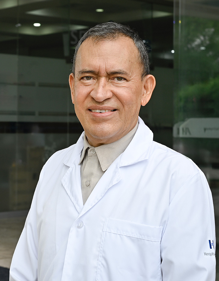 Es la foto de perfil de un médico general en Villahermosa de Hospital Angeles. Tiene puesto una bata blanca y una camisa color beige oscuro. Tiene el cabello grisaseo y ojos café claro. Está sonriendo ligeramente.
