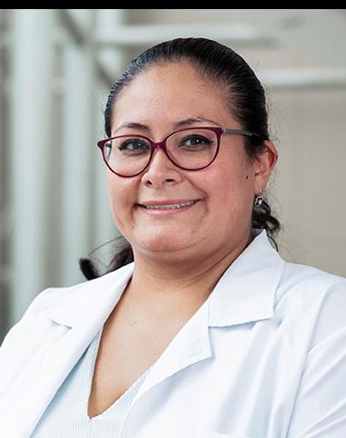 Es la foto de perfil de una ginecóloga y obstetra en Huixquilucan de Hospital Angeles Lomas. Usa una bata blanca con una blusa blanca, tiene lentes con marco rojo, el cabello recogido y está sonriendo.