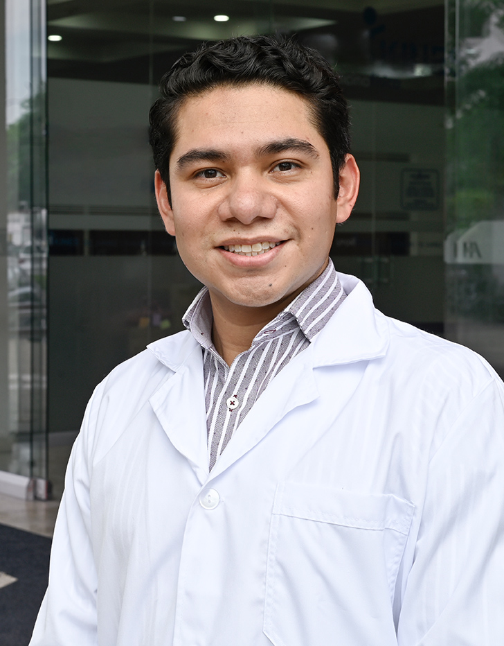 Es la foto de perfil de un oncólogo médico en Villahermosa de Hospital Angeles Villahermosa. Usa una bata blanca con una camisa color gris y líneas blancas. Tiene el cabello café oscuro y ondulado. Está sonriendo.