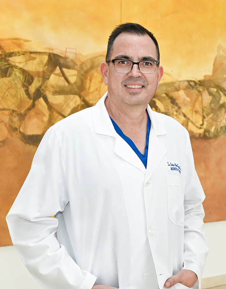 Es la foto de perfil de un ginecólogo y obstetra en Ciudad Juárez de Hospital Angeles Ciudad Juárez. Usa bata blanca, playera azul y lentes.