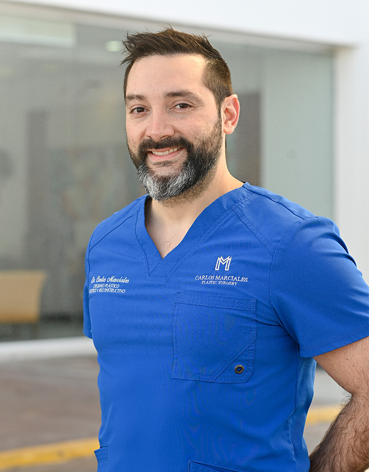 Es la foto de perfil de un cirujano plástico en Tijuana de Hospital Angeles Tijuana. Usa un uniforme médico de color azul. Tiene los brazos cruzados por detrás. Tiene el cabello café y la barba de color café oscuro con blanco.
