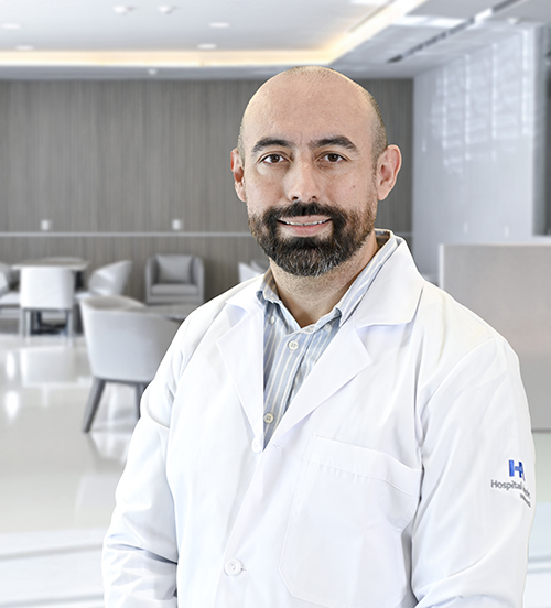 Es la foto de perfil de un cirujano neurológico y especialista en terapia endovascular neurológica en San Pedro Garza García de Hospital Angeles Valle Oriente. Usa una bata blanca con una camisa de cuadros azul, rojo y blanco. Está serio.