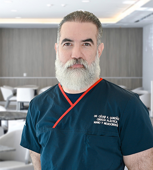 Es la foto de perfil de un cirujano plástico en San Pedro Garza García de Hospital Angeles Valle Oriente. Usa un uniforme médico color azul marino con bordes de color rojo. Tiene los brazos cruzados por detrás. Tiene el cabello y barba color gris claro.