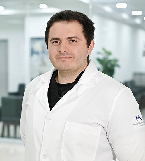 Es la foto de perfil de un ortopedista y traumatólogo en Ciudad de México de Hospital Angeles Acoxpa. Usa una bata blanca con un uniforme médico color negro. Tiene el cabello color café oscuro. Tiene los barzos cruzados por detrás.