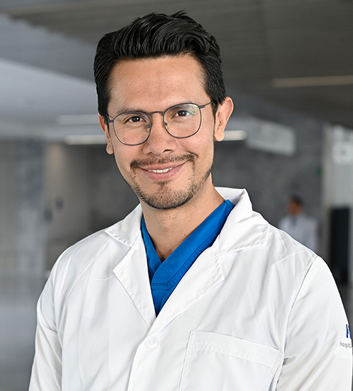 Es la foto de perfil de un cirujano maxilofacial en Querétaro de Hospital Angeles Centro-sur. Usa una bata blanca con la marca de Hospital Angeles. Tiene puesto un uniforme médico color azul. Tiene el cabello cafe oscuro y usa lentes.