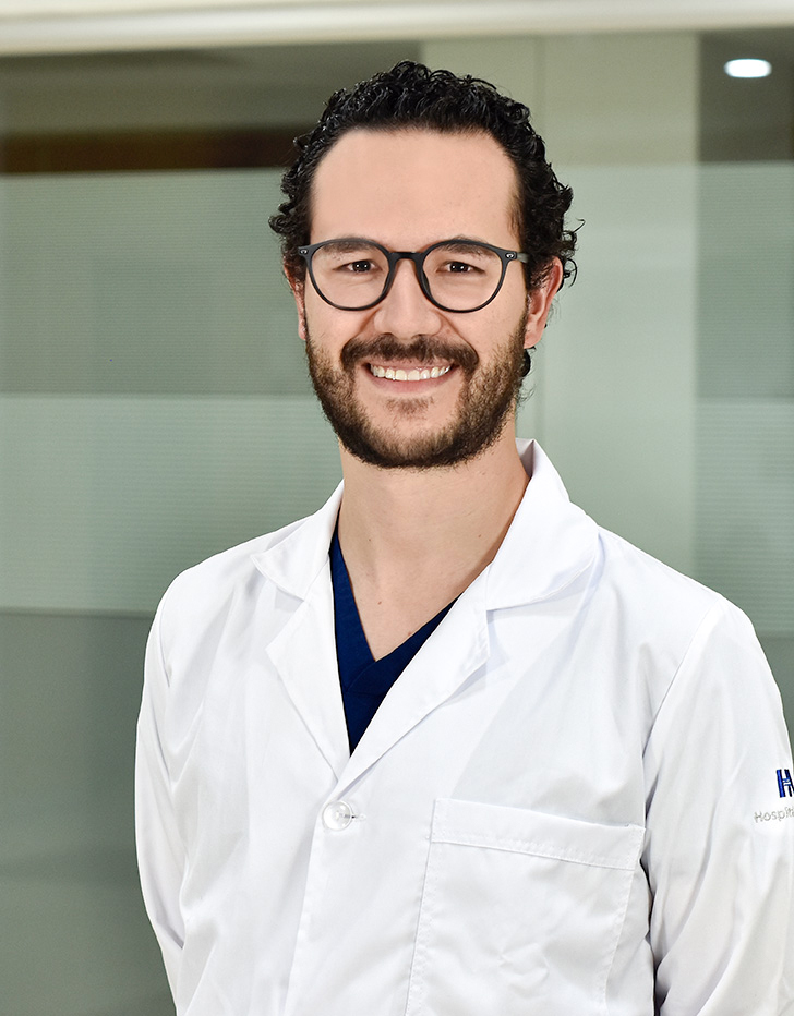 Es la foto de perfil de un cardiólogo y cardiólogo intervencionista en Ciudad de México de Hospital Angeles Mocel. Usa una bata abotonada color blanco con un uniforme color azul. Tiene barba y cabello color café oscuro. Usa lentes.
